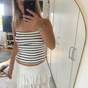 Topp - Säljer en vit bandeau tubtopp med svarta horisontella ränder. Toppen är ribbad och sitter tight mot kroppen, perfekt för varma dagar. Den är ärmlös och har en enkel, stilren look som passar till många outfits.
