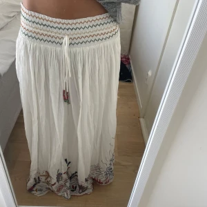 Vit långkjol med broderier och snörning - Superfin vit långkjol med broderade detaljer i färg längs nederkanten och smockad midja med snörning. Kjolen har vågiga kanter och broderier med havstema i rött, blått, grönt och gult.