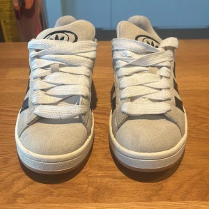 Adidas Campus sneakers i ljusgrå mocka - Snygga Adidas Campus sneakers i ljusgrå mocka med klassiska svarta ränder på sidorna och vit sula. Skorna har rund tå, platt sula och breda vita snören. Adidas logga syns på plös och häl. Perfekta för dig som gillar retrostil och streetwear. Använda ett fåtal gånger. 
