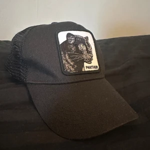 Svart Panther Keps - Cool svart keps med mesh på sidorna och en broderad patch framtill med en panter och texten 'Panther'. Perfekt för dig som vill sticka ut med en unik detalj. Justerbar baktill för bästa passform. Nypris: 400kr