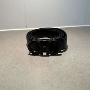 Svart skärp från Ferragamo i skinn - Snyggt svart bälte från Ferragamo i äkta skinn med en ikonisk dubbelringad metallspänne. Bältet har en stilren och modern look, perfekt för att lyfta vilken outfit som helst. Spännet har Ferragamo-loggan ingraverad för en exklusiv touch.