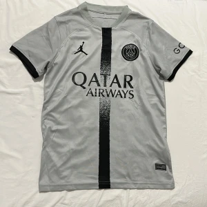 PSG grå fotbollströja Jordan x Nike - Snygg grå PSG fotbollströja med svart vertikal rand och svarta detaljer på ärmsluten. Tröjan har Paris Saint-Germain-logga, Jordan-logga och Qatar Airways-tryck på bröstet. Tillverkad i lätt och ventilerande material, perfekt för match eller träning.