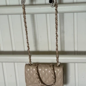 Beige quiltad axelväska med kedja - Snygg beige axelväska i quiltad design med guldfärgad kedja och läderdetaljer. Väskan har ett lock med lås och två små fack varav ett med med dragkedja. Perfekt storlek för mobil och småsaker, 18 cm bred. Stilren och trendig accessoar.