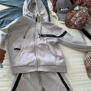 Ljusgrå dress från BOSS med dragkedja - Säljer en stilren ljusgrå hoodie från BOSS med hel dragkedja, svarta detaljer och snörning i huvan. Tröjan har fickor med svarta dragkedjor och en svart rand med BOSS-logga på bröstet. Perfekt för en chill och sportig look.