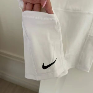 Vita träningstights-shorts från Nike - Snygga vita träningstights-shorts från Nike med hög midja och praktiska sidfickor. De är tillverkade i ett stretchigt syntetmaterial som sitter tight och bekvämt. Perfekta för gymmet eller löprundan, med klassisk svart Nike-logga på benet. Helt oanvänd!