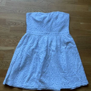 Vit axelbandslös spetsklänning - Superfin vit axelbandslös kortklänning med vackert spetsmönster och markerad midja. Klänningen har en söt, lätt utställd kjol och dragkedja bak för enkel påklädning.