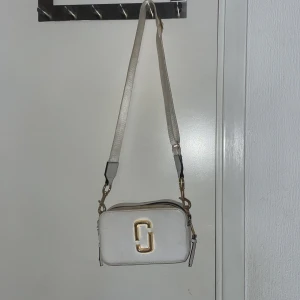 Vit axelväska från Marc Jacobs - Snygg vit axelväska från Marc Jacobs med gulddetaljer och justerbar axelrem. Väskan har en praktisk dragkedja och en stilren design med logotyp framtill. Perfekt för att bära dina nödvändigheter med stil.