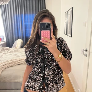 Svart paljettopp från Gina Tricot - Säljer en glittrig svart topp från Gina Tricot, täckt med paljetter och dekorerad med svarta rosetter framtill. Toppen har korta ärmar och en elegant design som passar perfekt för festliga tillfällen.💖💞