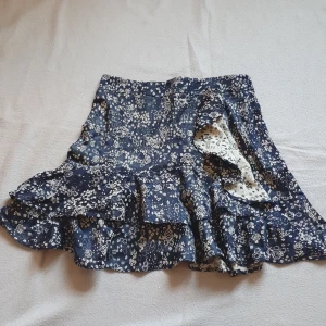 Blommig kjol från Zara - Söt blommig kjol från Zara i svart (eller mörkblått??) med guldiga blommor. Kjolen har volanger och en omlott-design som ger en fin rörelse. Sparsamt använd för den är så fiiin 🖤