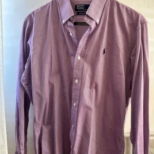 Lila skjorta från Polo - Säljer en stilren lila skjorta från Polo i 100% bomull. Skjortan har en klassisk krage med knappar och en broderad logotyp på bröstet. Perfekt för en snygg och avslappnad look.