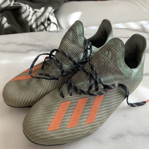 Adidas fotbollsskor i grönt och orange - Snygga Adidas fotbollsskor i en cool grön färg med orange detaljer. Skorna har snörning och en rund tå, perfekt för fotbollsplanen. De har en modern design med de klassiska tre ränderna på sidan. Lite jord på dobbarna men de går att tvätta bort lätt