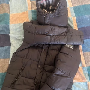 Svart dunjacka från Burberry - Snygg svart dunjacka från Burberry med avtagbar huva och dragkedja. Jackan har ett klassiskt rutigt foder och praktiska fickor med dragkedja. Perfekt för kyliga dagar.