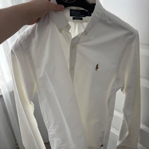Vit skjorta från Ralph Lauren - Säljer en klassisk vit skjorta från Ralph Lauren med broderad logga på bröstet. Skjortan har långa ärmar och knappar framtill. Perfekt för en stilren look.