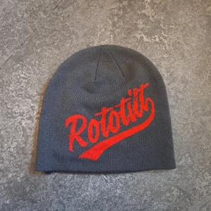 Grå mössa med röd text - Snygg grå mössa med röd text från Rototilt. Perfekt för att hålla värmen under kyliga dagar. Den har en enkel design med en liten etikett på sidan.