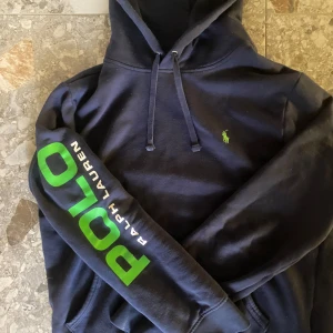 Mörkblå hoodie från Ralph Lauren - Säljer en snygg mörkblå hoodie från Ralph Lauren med grönt POLO-tryck på ärmen och en liten grön logga på bröstet. Perfekt för en avslappnad stil. Huvan har dragsko för justering.