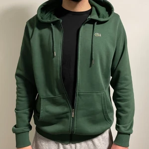 Grön huvtröja från Lacoste - Säljer en ÄKTA, snygg grön huvtröja från Lacoste med dragkedja och justerbar huva. Köpt från Milano för 5 månader sedan. Koftan har långa ärmar och ribbade muddar för extra komfort! Koftans storlek är XL fast eftersom det är bomull har den perfekta passformen för L också. 