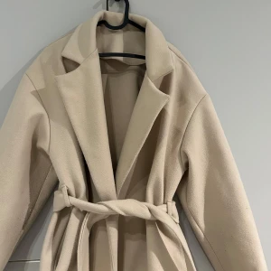 Beige kappa med bälte - Elegant beige kappa med bälte i midjan. Perfekt för kyligare dagar med sin stilrena design och långa ärmar. Kappan har en klassisk krage och är tillverkad i ett mjukt material.