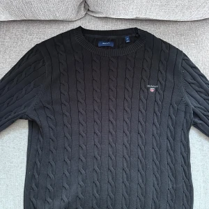 Svart stickad tröja från Gant - Säljer en stilren svart stickad tröja från Gant med kabelstickat mönster. Tröjan har rund hals och långa ärmar, perfekt för kyligare dagar. Diskret logga på bröstet. Passar både till vardags och mer uppklädda tillfällen.