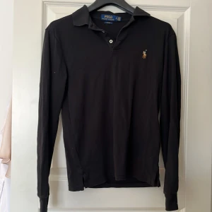 Svart pikétröja från Ralph Lauren - Säljer en stilren svart pikétröja från Ralph Lauren i slim fit. Tröjan har långa ärmar och en klassisk krage med knappar. Den ikoniska broderade logotypen syns på bröstet. Perfekt för en elegant och avslappnad look. Notera skadan vid knapp-hålen