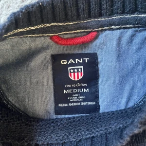 Blå stickad tröja från GANT - Säljer en blå stickad tröja från GANT med ett stort amerikanskt flaggmotiv på framsidan. Tröjan har långa ärmar och en rund halsringning. Perfekt för en avslappnad stil med en touch av amerikansk inspiration. Priset kan diskuteras! Men går inte under 200 kr. 🤝
