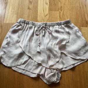 Silkesaktigt Beige shorts med knytband XS - Säljer ett par luftiga beige shorts med elastisk midja och knytband med tofsdetalj. Perfekta för varma sommardagar. De har en avslappnad passform och är gjorda i ett lätt silkesmaterial. Bra skick! Tags bortklippta. 