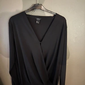 Svart omlottopp från Lindex - Säljer en elegant svart omlottopp från Lindex. Toppen har en v-ringad design och långa ärmar, perfekt för en stilren look. Passar bra till både jeans och kjol. Perfekt för dig som vill ha något enkelt men snyggt i garderoben.