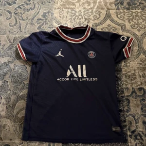 Mörkblå PSG fotbollströja - Säljer en mörkblå PSG fotbollströja från Jordan med vita och röda detaljer. Tröjan har korta ärmar och klubbens emblem på bröstet. Köpt på Zalando