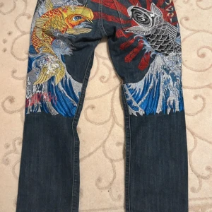 Japansk denim - Unika jeans från Sugoi med detaljerat japanskt broderi av karpar och vågor i färgerna röd, blå och gul. Jeansen har en klassisk femficksdesign och är tillverkade i bomull. Midja 42 cm längd 100cm benöpning 20cm