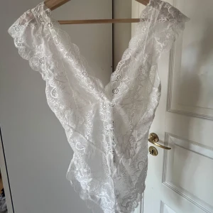 Vit spetsbody - Säljer en elegant vit spetsbody med djup v-ringning och vackra spetsdetaljer. Perfekt för en romantisk look eller som en del av en festoutfit. Den är ärmlös och har en fin passform som framhäver figuren. Aldrig använd eller provad
