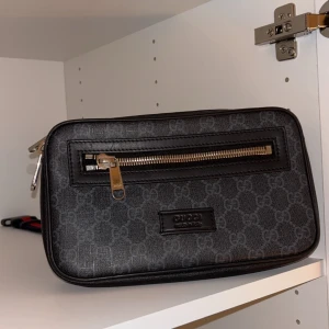 Svart axelväska från Gucci - Snygg svart axelväska från Gucci med ett diskret mönster och silverfärgade dragkedjor. Väskan har en justerbar rem i rött och blått, perfekt för en stilren look. Den har flera fack för enkel organisering. Helt äkta. Pris kan diskuteras.