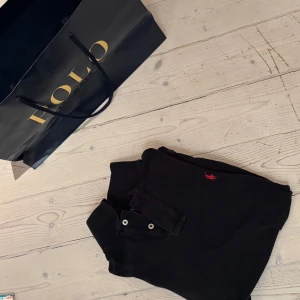 Svart pikétröja från Polo Ralph Lauren - Säljer en klassisk svart pikétröja från Polo Ralph Lauren med den ikoniska röda loggan broderad på bröstet. Tröjan har en krage med knappar och är perfekt för en stilren look. Passar både till vardags och mer uppklädda tillfällen.