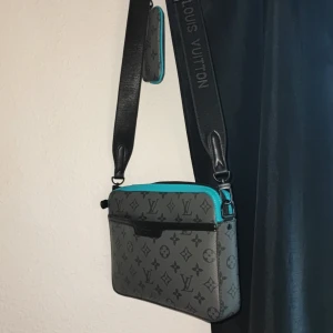 Trio messenger från Louis Vuitton - Säljer en snygg grå axelväska från Louis Vuitton med det klassiska monogrammönstret. Väskan har svarta detaljer och en dragkedja för säker förvaring. Den har också en justerbar axelrem för bekväm användning. Perfekt för dig som vill ha en stilren och lyxig väska.