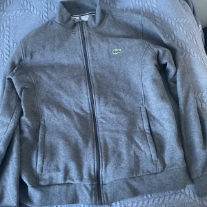 Lacoste Zip - Säljer en stilren grå ziphoodie från Lacoste med dragkedja och ståkrage. Jackan har långa ärmar och två fickor framtill. Perfekt för en avslappnad look. Det är fransk storlek vilket betyder att de är väldigt små. Står 6 vilket motsvarar L. 