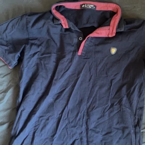 Mörkblå kortärmad pikétröja från B. Jones - Snygg mörkblå pikétröja från B. Jones med korta ärmar och en kontrasterande rosa krage. Tröjan har en liten broderad logga på bröstet och knappar vid halsen. Perfekt för en avslappnad stil.