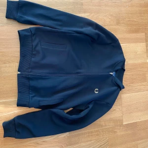 Svart bomberjacka från Fred Perry - Snygg svart bomberjacka från Fred Perry med dragkedja och ribbade muddar. Jackan har en broderad logga på bröstet och två fickor framtill. Perfekt för en stilren look.