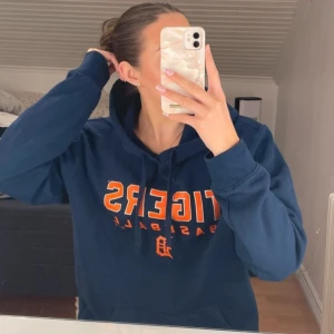 Blå hoodie med Tigers-tryck - Säljer en blå hoodie med texten 'Tigers Baseball' i orange på framsidan. Tröjan har en klassisk känguruficka och justerbar huva med dragsko. Perfekt för en avslappnad stil.