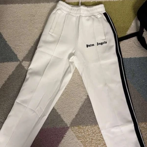 Vita byxor från Palm Angels (äkta) - Snygga vita byxor från Palm Angels med svarta ränder längs sidorna. Byxorna har en modern design med dragkedja vid fickorna och en bekväm passform. Perfekta för en stilren look. Med prislapp aldrig använd 