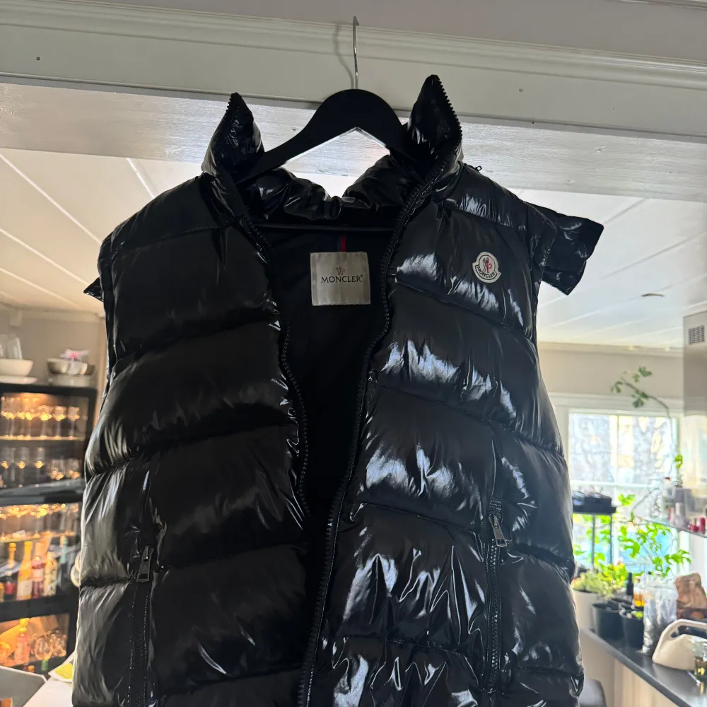 Snygg svart dunväst från Moncler med glansig finish och dragkedja framtill. Perfekt för att hålla värmen med stil. Västen har en hög krage och Moncler-logga på bröstet.. Takit.