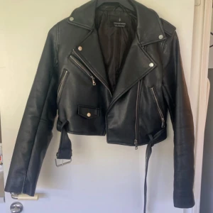 Svart skinnjacka från Stradivarius - Snygg svart skinnjacka från Stradivarius med en cool croppad design. Jackan har dragkedjor och knappar som detaljer, vilket ger den en edgy look. Perfekt för att lyfta vilken outfit som helst. Tyvärr är den lite sliten vid armen men det är inget som märks av när man väl har på sig den. Det är även lite vit färg på den som man ser på bilden. 