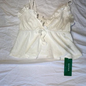 Vit topp med volangdetaljer - Säljer en söt vit topp med volangdetaljer och knytning framtill. Toppen har smala axelband och en lätt utsvängd passform. Perfekt för varma dagar!