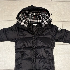 Burberry pufferjacket - Snygg svart dunjacka från Burberry med ett stilrent rutigt foder i svart, vitt och rött. Jackan har en praktisk dragkedja och knappar framtill samt en huva för extra värme. Perfekt för kyliga dagar!                                                                               Vill helst byte mot något mer somrigt kanske en sommarjacka eller väst eller skor typ kom med förslag så svarar jag inom kort!!!