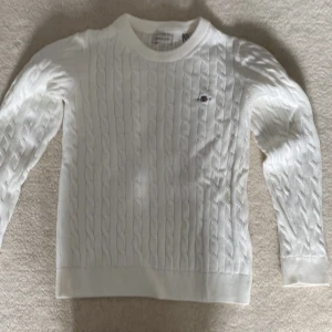 Vit stickad tröja från GANT - Säljer en stilren vit stickad tröja från GANT med klassiskt flätmönster. Tröjan har långa ärmar och en rund halsringning.  Den är mjuk och bekväm, idealisk för både vardag och speciella tillfällen. Helt oanvänd.