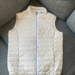 Beige dunväst från Slam - Snygg beige dunväst från Slam i storlek M. Västen har en dragkedja framtill och en stilren design som passar perfekt för kyligare dagar. Perfekt för lager-på-lager-stil.