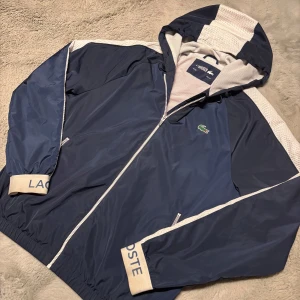 Blå och röd vindjacka från Lacoste - Snygg vindjacka från Lacoste i blått och rött med vita detaljer. Jackan har en dragkedja framtill och en huva. Elastiska muddar med Lacoste-logga på ärmsluten. Perfekt för vår och höst.