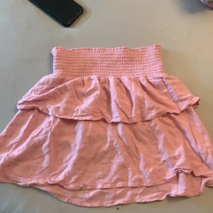 Rosa kjol med volang - Söt rosa kjol med volangdetaljer och en smockad midja som ger en bekväm passform. Perfekt för varma dagar och en avslappnad stil. Kjolen har en kort längd och är gjord i ett lätt material.