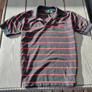Grå och röd randig pikétröja från Fred Perry - Säljer en klassisk svart pikétröja från Fred Perry med röda ränder. Tröjan är tillverkad i 100% bomull och har korta ärmar samt en broderad logga på bröstet. Perfekt för en stilren look!