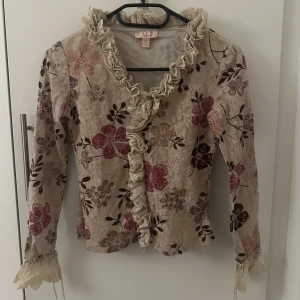 Blommig spetsblus med volangdetaljer - Säljer en romantisk blommig spetsblus med volangdetaljer. Storlek 36, men passa som 34. Blusen har en beige bas med blommönster i rosa och bruna nyanser. Den har långa ärmar och volanger längs kragen och ärmsluten. Perfekt för en bohemisk look!