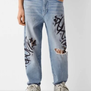 Graffitimönstrade jeans från Bershka - Säljer ett par coola ljusblå jeans från Bershka med graffitimönster och slitna detaljer. Jeansen har en normal passform och är perfekta för en avslappnad stil. De har klassisk femficksdesign och knappgylf.