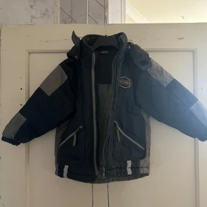 Grå och svart jacka från Race Team - Säljer en grå och svart jacka från Race Team med huva och dragkedja. Jackan har långa ärmar och flera fickor med dragkedjor. Perfekt för kyligare dagar med sin fodrade insida.