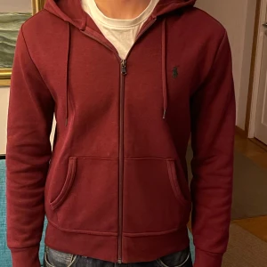 Vinröd hoodie från Ralph Lauren - Säljer en snygg vinröd hoodie från Ralph Lauren med dragkedja och huva. Perfek till våren eller sommaren. I ett stretch tyg. I använt skick men inga större defekter. Jag är 181 på bilden och den  är något kort. Tveka inte på höra av dig vid frågor!